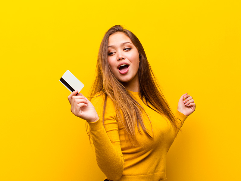 Imagem com fundo amarelo e uma mulher no centro, segurando um cartão de crédito e com expressão de felicidade por causa do valor da lipoescultura.