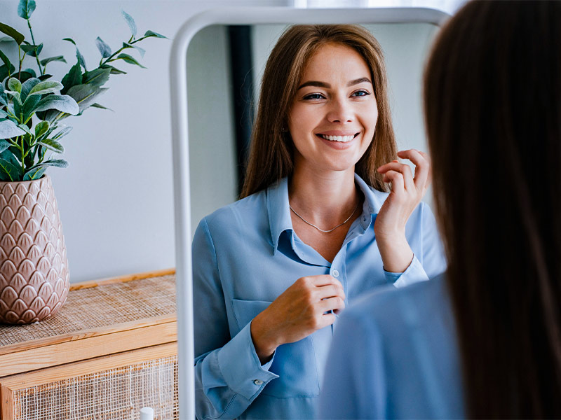 Mulher de camisa azul sorrindo enquanto se olha no espelho, completamente feliz depois de feito uma plástica