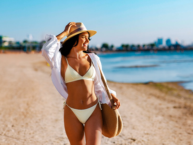 Mulher na praia, segurando o chapéu, olhando para o mar sorrindo, depois da redução mamária