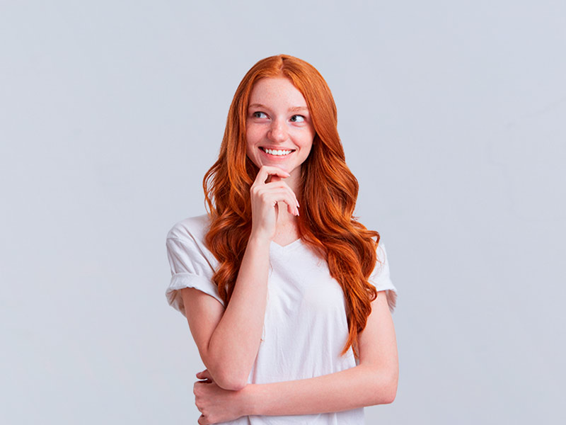 Menina ruiva de camiseta branca, sorrindo com a mão no queixo, olhando para o lado direito, pensando a partir de qual idade pode fazer a mastopexia