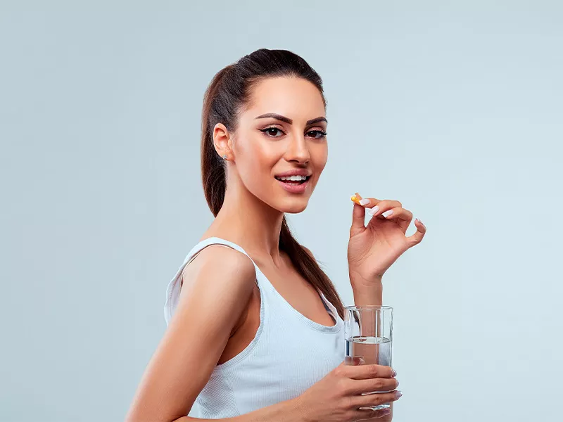 Tomar colágeno com ácido hialurônico é seguro? Imagem de uma mulher segurando uma cápsula de colágeno com ácido hialurônico e um copo d'água