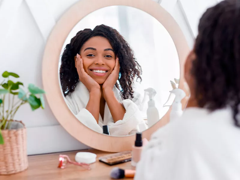 Você sabe para que serve o ativo? Imagem de uma mulher se olhando no espelho com expressão de felicidade após saber os benefícios do ácido hialurônico para o rosto