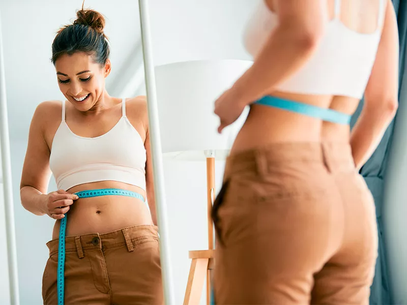 Quantos centímetros serão retirados? Mulher medindo a cintura se olhando no espelho, ela está feliz por saber que poderá fazer Abdominoplastia.