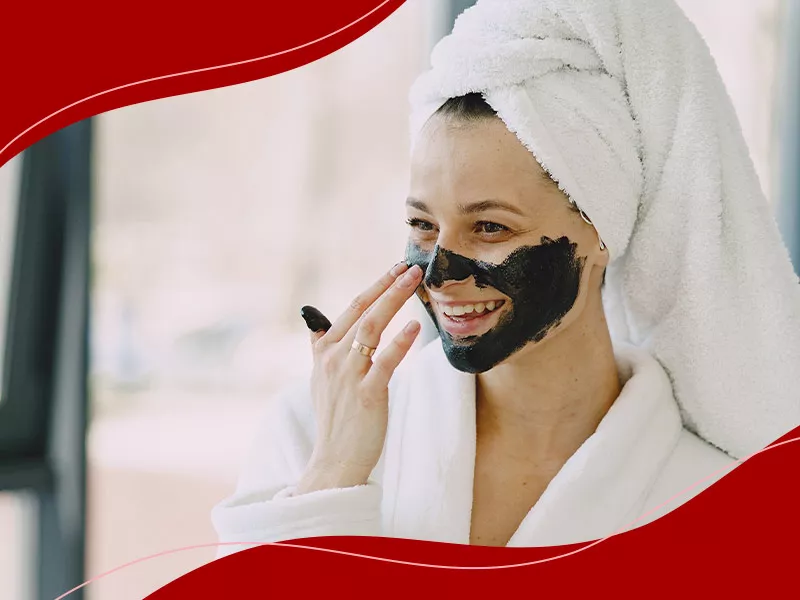 Mulher de roupão e toalha branca no cabelo, passando máscara de carvão no rosto